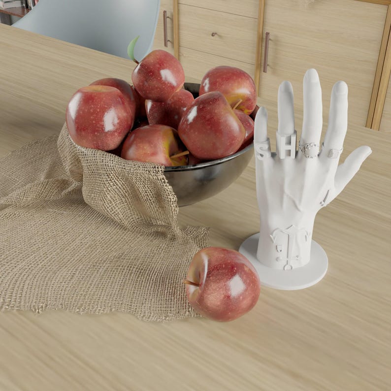 May include: A still life featuring a bowl of red apples, a white hand-shaped ring holder with rings, and a single apple. The apples are in a metal bowl on a burlap cloth, set on a wooden surface.