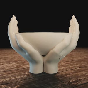 May include: A white decorative bowl in the shape of two hands cupping a round vessel. The hands are sculpted with detailed fingers and a smooth finish. The bowl is set against a dark background and a wooden surface.