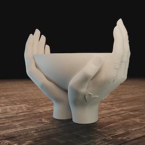 May include: A white decorative bowl held by two hands. The hands are sculpted with intricate details, including rings and a heart-shaped design. The bowl is a smooth, rounded shape, perfect for holding small items.