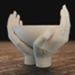 May include: A white, open-palmed hand sculpture with a small, bowl-shaped space between the hands. The sculpture is supported by two cylindrical legs. The hands and bowl are smooth, and the sculpture is set against a dark background.