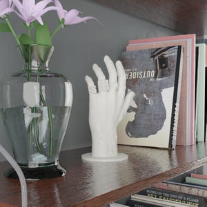 May include: A white hand sculpture with rings spelling "SH". A clear glass vase holds pink flowers. Books with the title "The Outsider" are on a wooden shelf. The shelf is against a gray wall.