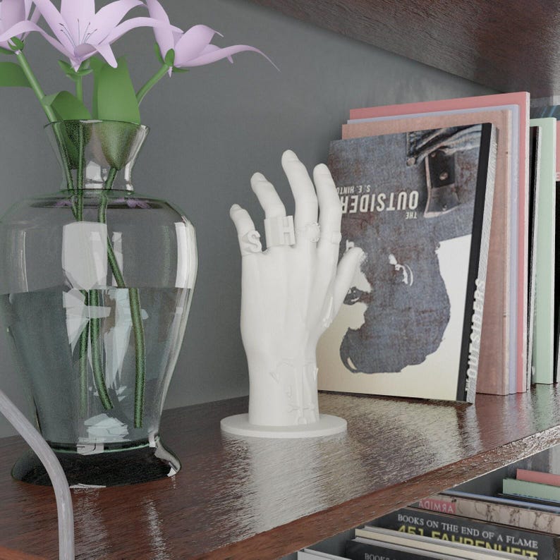 May include: A white hand sculpture with rings spelling "SH". A clear glass vase holds pink flowers. Books with the title "The Outsider" are on a wooden shelf. The shelf is against a gray wall.