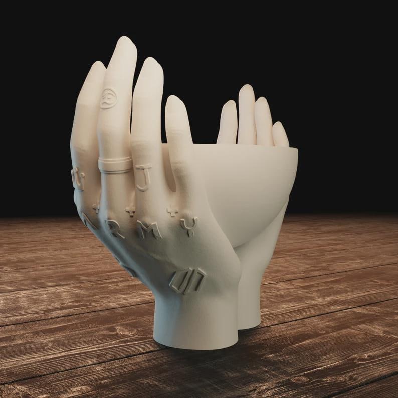May include: A white ceramic sculpture of two hands cupped together, forming a bowl. The fingers are detailed with rings and tattoos spelling "ARMY". The sculpture is set against a dark background on a wooden surface.
