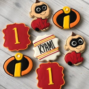 May include: Set of six decorated sugar cookies featuring the character 'Baby Jack-Jack' from the movie 'Incredibles 2'. The cookies are decorated with red, yellow, black, and white icing. Two cookies feature the number '1' in yellow icing on a red background. One cookie features the name 'KYAMI' in black icing on a white background with yellow and orange stripes. Two cookies feature the 'Incredibles' symbol in black, yellow, and orange icing. One cookie features 'Baby Jack-Jack' in red and white icing.