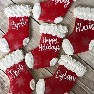 May include: Red Christmas stocking cookies with white frosting trim and snowflake details. Each cookie is personalized with a name or the phrase "Happy Holidays" in white icing. The cookies are arranged on a wooden surface.