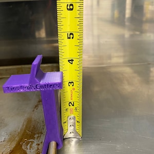 Puede incluir: Herramienta de plástico púrpura etiquetada como "Tiny Owls Cutters" medida con una cinta métrica amarilla. La herramienta tiene 7,6 cm de altura.