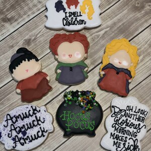 Puede incluir: Conjunto de nueve galletas de azúcar con temática de Halloween decoradas con glaseado de colores. Las galletas representan brujas, un caldero y texto que dice "I Smell Children", "Amuck", "Hocus Pocus" y "Oh Look! Another Glorious Morning Makes Me Sick".