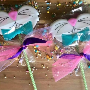Puede incluir: Dos galletas con forma de conejo en palos, decoradas con glaseado blanco, ojos negros, narices rosas y lazos de color turquesa. Las galletas están envueltas en celofán transparente con lazos de tul rosa y morado y espolvoreadas con coloridos.