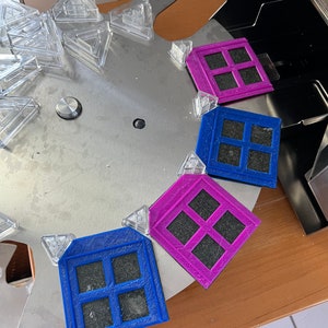 Puede incluir: Un surtido de formas geométricas y objetos cuadrados coloridos con diseños de ventanas. Los objetos son en tonos de azul y morado, con un interior gris oscuro. También hay piezas triangulares transparentes.