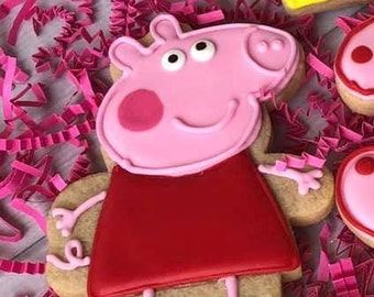 May include: A pink and red gingerbread cookie decorated with icing to look like Peppa Pig. The cookie is on a pink and white background.