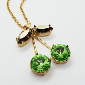 May include: A gold chain necklace with a green and black crystal cherry pendant. The pendant features two round green crystals and a black oval crystal.