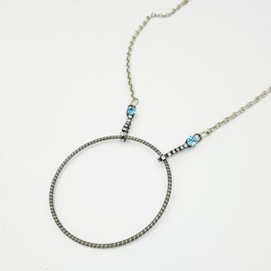 May include: A silver chain necklace with a circular pendant made of a coiled metal wire. The pendant is attached to the chain with two strands of rhinestones and blue gemstones.