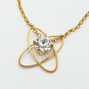 May include: A gold chain necklace with a clear crystal pendant. The pendant is set in a gold frame that resembles a stylized atom.