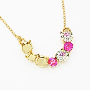 May include: A gold-colored necklace with a delicate chain. The necklace features a series of round, bezel-set gemstones in shades of pink and clear, arranged in a curved design. The necklace is set against a white background.