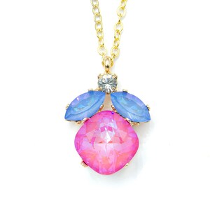 May include: A gold-toned necklace featuring a pendant with a large, pink, square-cut crystal at the center, flanked by two blue, leaf-shaped crystals. A small, clear crystal sits above the pink crystal. The chain is also gold.