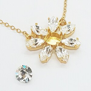 May include: A gold-colored necklace with a flower-shaped pendant. The pendant features clear, marquise-cut crystals arranged around a central setting. A single, round, clear crystal sits nearby. The necklace is on a white background.