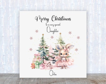 Puede incluir: Una tarjeta de Navidad con una ilustración de acuarela de un conejito con un tutú rosa y un lazo, de pie frente a un bosque nevado con árboles de Navidad y regalos. La tarjeta dice "Feliz Navidad a una hija muy especial" y está personalizada con el nombre "Orla".