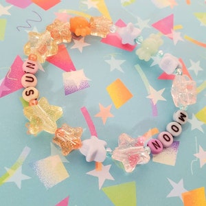 Pu&ograve; includere: Un braccialetto di perline colorate con le parole "MOON" e "SOON" scritte in lettere bianche. Le perline sono un mix di colori pastello e forme a stella.