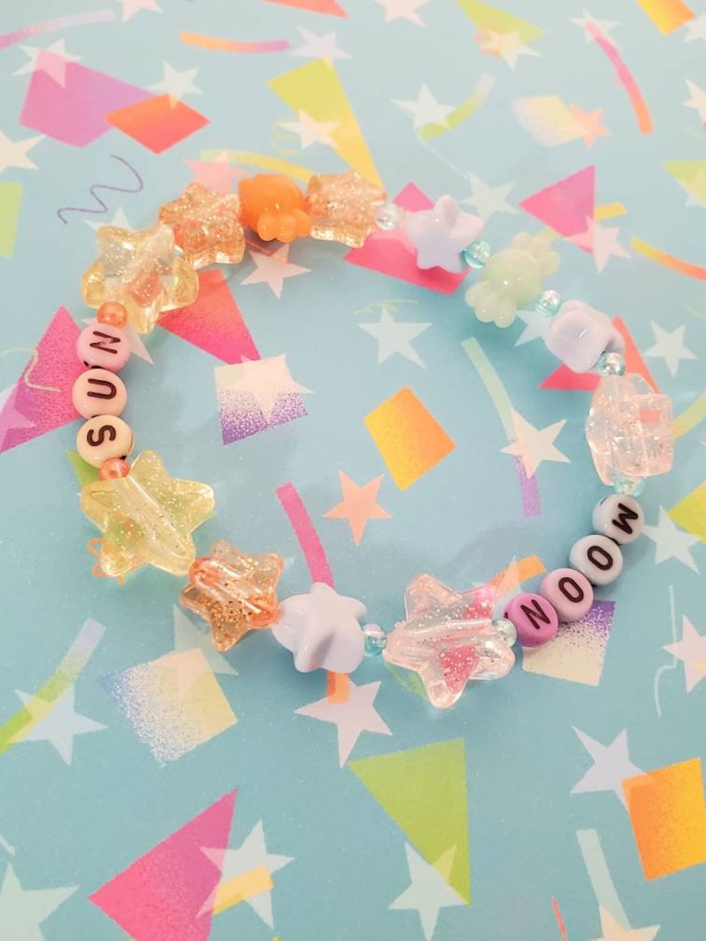 Pu&ograve; includere: Un braccialetto di perline colorate con le parole "MOON" e "SOON" scritte in lettere bianche. Le perline sono un mix di colori pastello e forme a stella.