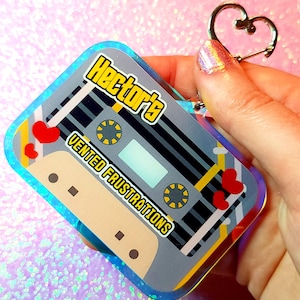 May include: A rectangular keychain featuring a cassette tape design. The keychain has a gray background with yellow and red accents, and the text "Hector's Vented Frustrations". It includes a heart-shaped clasp.