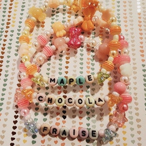 Pu&ograve; includere: Tre braccialetti colorati con perline con le parole "Maple", "Chocola" e "Fraises" scritte in lettere nere.