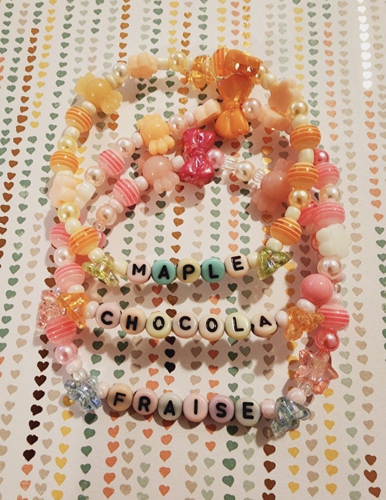 Pu&ograve; includere: Tre braccialetti colorati con perline con le parole "Maple", "Chocola" e "Fraises" scritte in lettere nere.