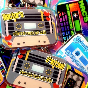 May include: A collection of colorful cassette tape-shaped keychains. One keychain features the text "Hector's" and "Vented Frustrations" on a gray background with red heart accents. Other keychains have various designs and text.