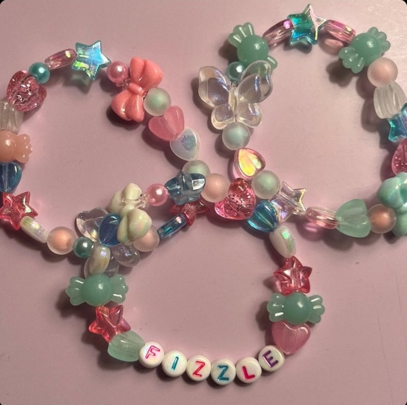 Pu&ograve; includere: Tre braccialetti di perline colorate con una variet&agrave; di ciondoli, tra cui stelle, cuori, farfalle e caramelle. I braccialetti compongono la parola "FIZZLE" in lettere bianche.