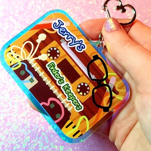 May include: A rectangular keychain featuring a cassette tape design with the text "Jerry's Finder's Keepers." The keychain has a heart-shaped clasp and a colorful, retro aesthetic with a blue border and glasses graphic.
