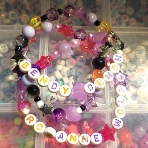 Pu&ograve; includere: Due braccialetti colorati con perline con le parole "Bendy Demon" e "Roxanne Wolf" scritte in lettere bianche con bordi dorati. I braccialetti sono realizzati con perline rosa, viola e bianche, oltre a ciondoli a forma di stella e cuore.