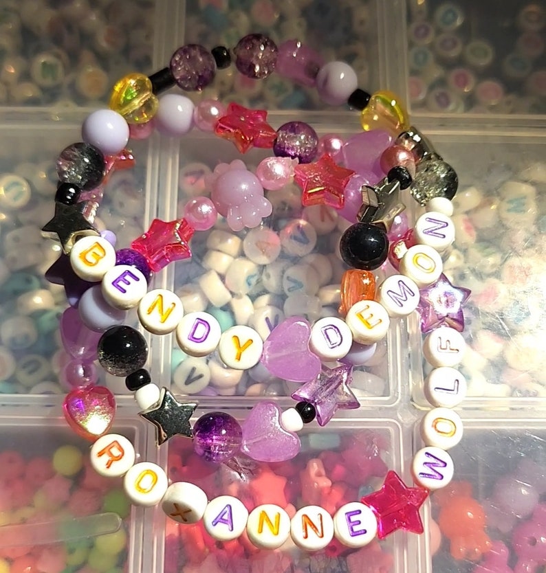 Pu&ograve; includere: Due braccialetti colorati con perline con le parole "Bendy Demon" e "Roxanne Wolf" scritte in lettere bianche con bordi dorati. I braccialetti sono realizzati con perline rosa, viola e bianche, oltre a ciondoli a forma di stella e cuore.