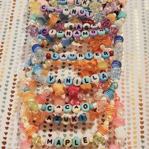 Pu&ograve; includere: Un mucchio di braccialetti colorati con perline, con diversi nomi scritti su di essi, tra cui "LK", "Coconut", "Sage", "Cinnamon", "Laurier", "Vanilla", "Cacao", "Azuki", "Maple", "Chocola" e "Fraise".