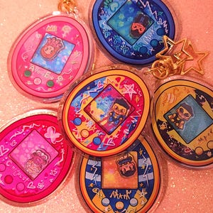 May include: A collection of colorful acrylic keychains in the shape of retro handheld electronic games. Each keychain features a unique design with cartoon characters and decorative elements. The keychains have gold-colored star-shaped clasps.