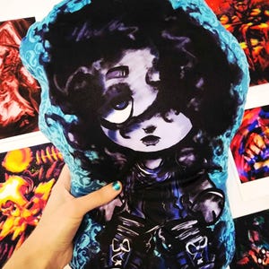 May include: A plush pillow featuring a stylized illustration of a girl with dark hair and large eyes. The pillow has a blue patterned border and is held against a background of artwork.