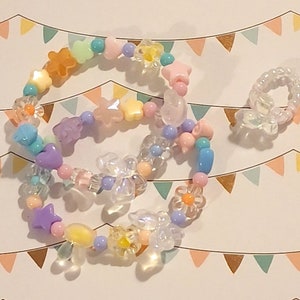 Pu&ograve; includere: Un braccialetto di perline colorate con perline rosa pastello, gialle, blu e viola. Le perline hanno la forma di stelle, fiori e cuori. Il braccialetto &egrave; su uno sfondo bianco con un motivo di triangoli color pastello.