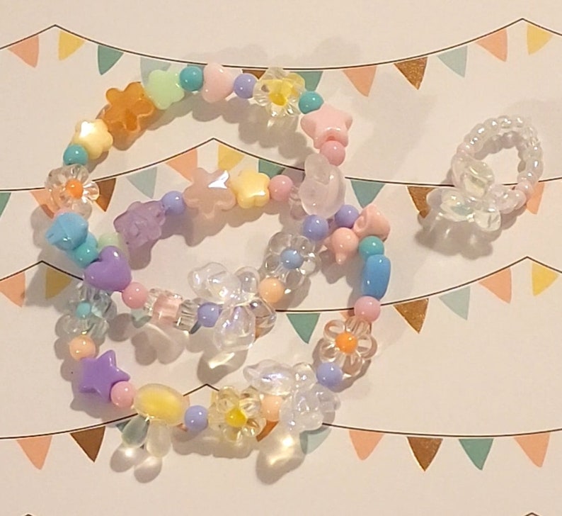 Pu&ograve; includere: Un braccialetto di perline colorate con perline rosa pastello, gialle, blu e viola. Le perline hanno la forma di stelle, fiori e cuori. Il braccialetto &egrave; su uno sfondo bianco con un motivo di triangoli color pastello.