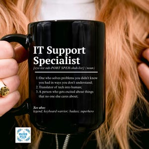 May include: Black ceramic mug with the text "IT Support Specialist" and a definition. The mug also includes the text "See also: legend; keyboard warrior; badass; superhero". The mug is held by a person.