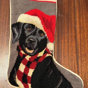 May include: A needlepoint Christmas stocking featuring a black Labrador retriever wearing a red and white Santa hat and a red, white, and black plaid scarf. The stocking has a cream-colored cuff and trim against a gray background.