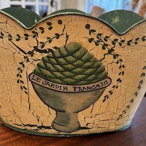 May include: A green and cream-colored metal planter with a scalloped rim. The planter has a distressed finish and features a painted design of a green artichoke in a pot with the text "LE JARDIN FRANCAIS".