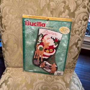 May include: A Bucilla felt applique kit for an 18-inch diagonal stocking featuring a Santa Claus, a reindeer, and forest animals. The packaging includes gold-plated needles and instructions for personalization with the name Lauren.