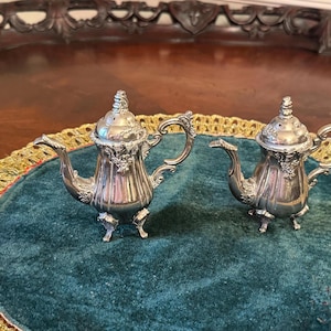 May include: Two silver teapot-shaped salt and pepper shakers with ornate details on a green velvet surface.