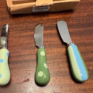 May include: Three metal knives with ceramic handles. The first handle is light green with blue polka dots, the second is medium green with light green polka dots, and the third is green with a vertical blue stripe. A cardboard box is visible above.