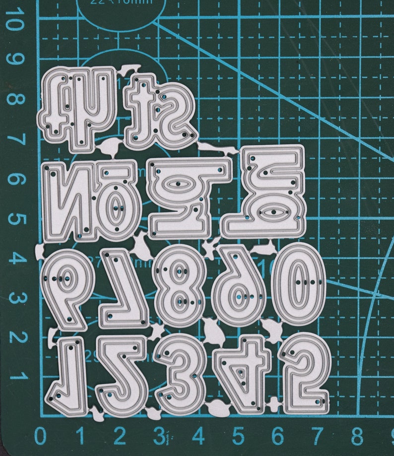 May include: A set of metal number dies, ranging from 0 to 9, plus additional numbers. The dies are silver-colored and have a rounded font. They are arranged on a green cutting mat with grid lines.