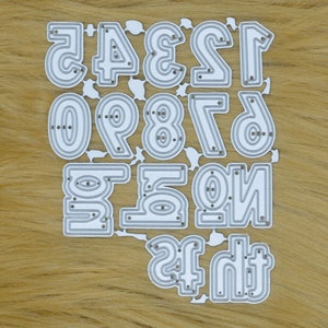 May include: A die-cut set of numbers 0-9 and the numbers 1-6 in reverse. The numbers are in a sans-serif font with a double-lined design. The die-cut is silver and sits on a light brown, textured surface.