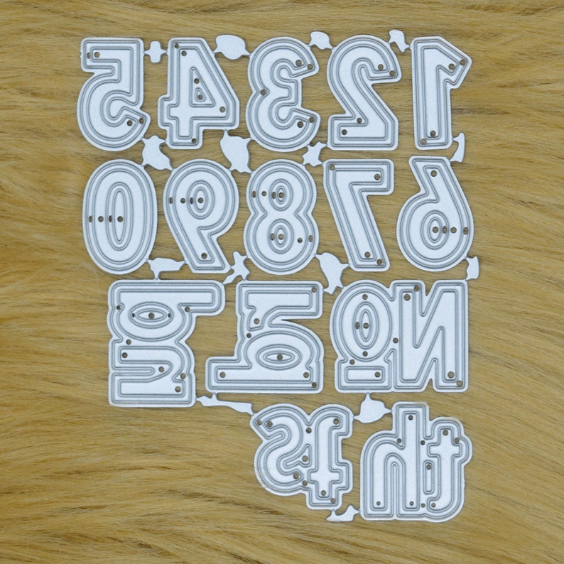 May include: A die-cut set of numbers 0-9 and the numbers 1-6 in reverse. The numbers are in a sans-serif font with a double-lined design. The die-cut is silver and sits on a light brown, textured surface.