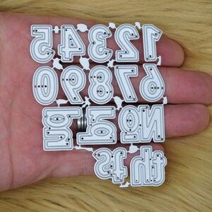 May include: A set of silver metal number dies, featuring the digits 0 through 9, plus the numbers 1 and 2. The dies are designed with a layered effect and small holes for detail. The numbers are held in a hand.