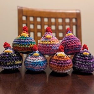 May include: Seven crocheted chick figurines in various colors, including purple, pink, orange, blue, and green. Each chick has a red beak and a red comb on its head.