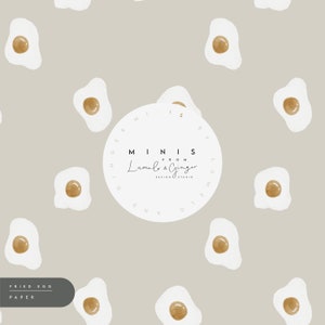 May include: A repeating pattern of watercolor fried eggs on a light gray background. Each egg has a white egg white and a golden yolk. The text "FRIED EGG PAPER" is at the bottom left. The design is from Lumel & Ginger.