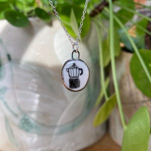 Op de afbeelding: Een zilveren ketting met een witte keramische hanger met een zwart-witte illustratie van een koffiepot.