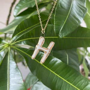 Könnte beinhalten: Eine goldene Kettenkette mit einem weißen Keramik-Anhänger in Form des Buchstabens H. Der Anhänger hat einen goldenen Umriss.